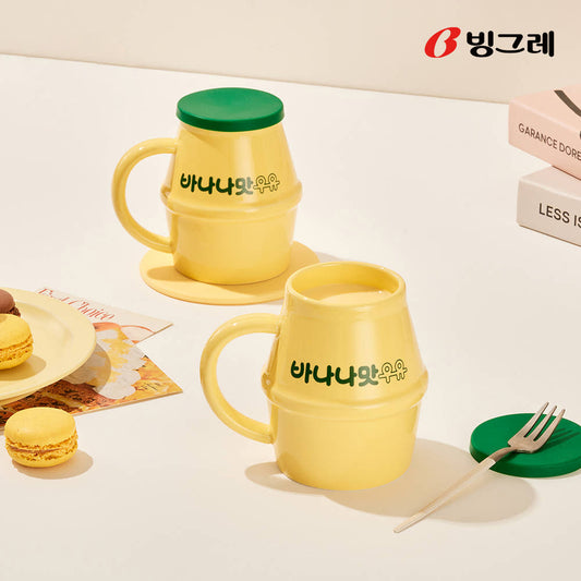 Two yellow Binggrae banana milk ceramic mugs with green silicone lids, styled in a bright setting with snacks.